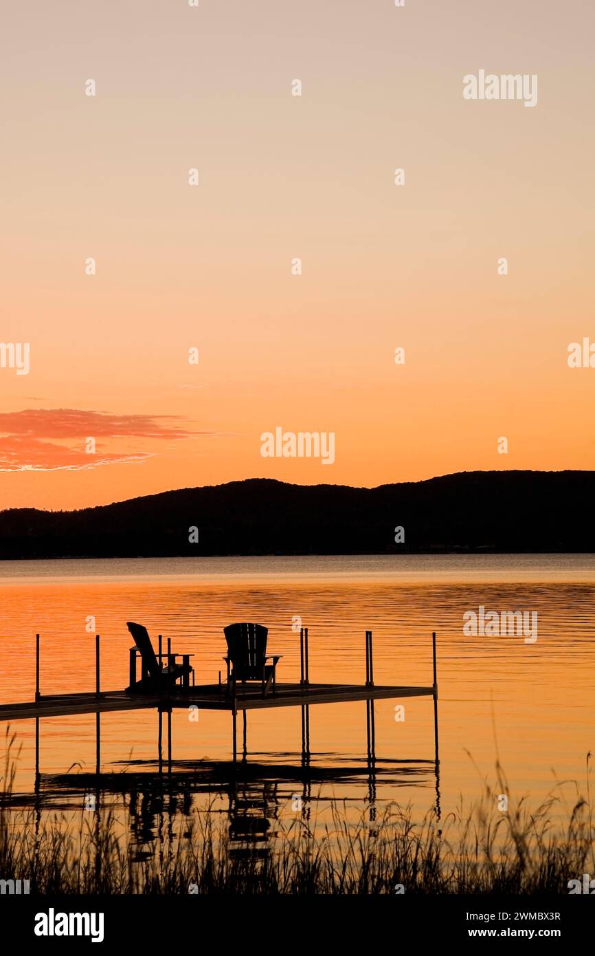 Silhouetted raised dock with Adirondack chairs over lake at sunset in ...