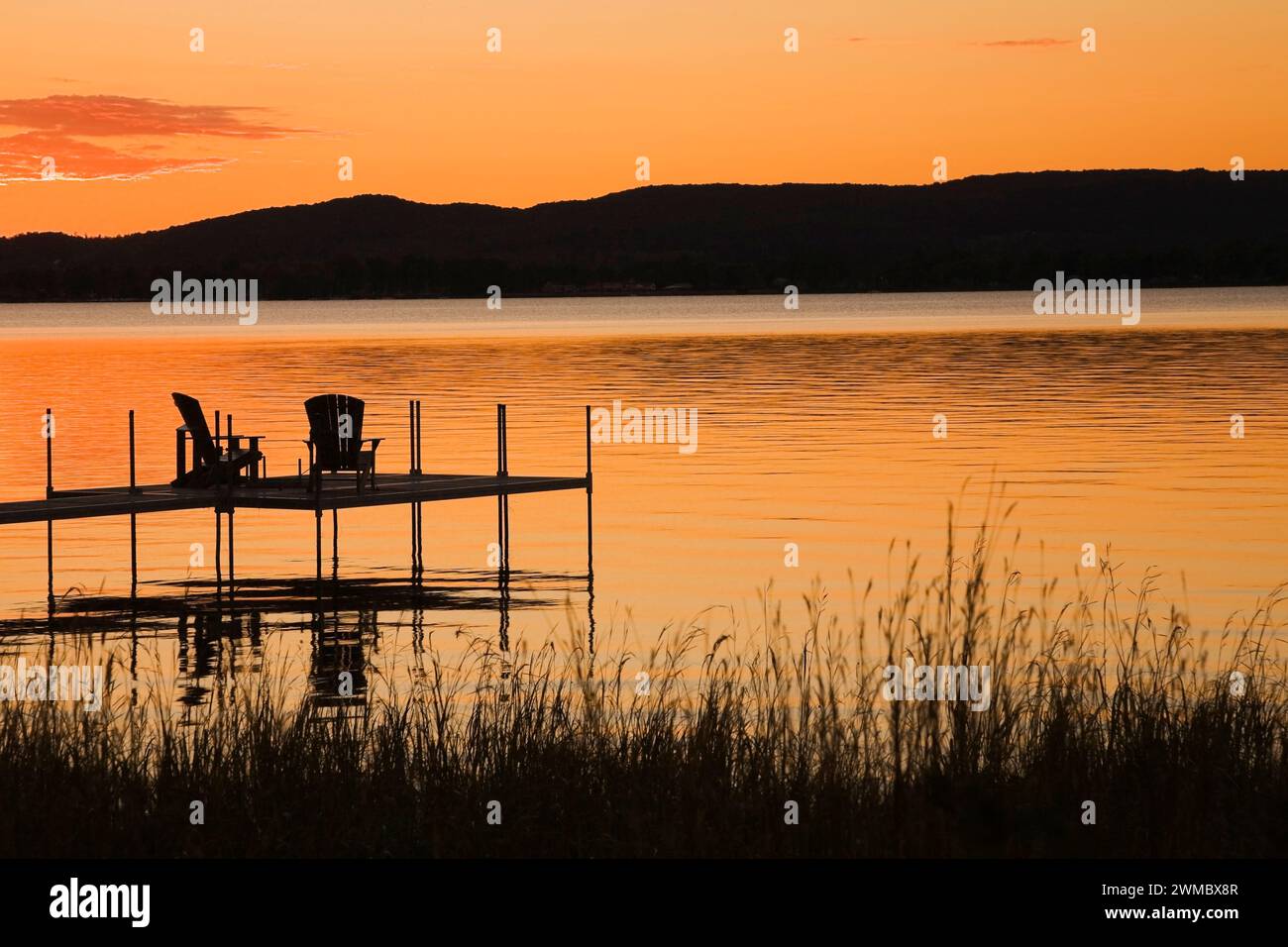 Silhouetted raised dock with Adirondack chairs over lake at sunset in