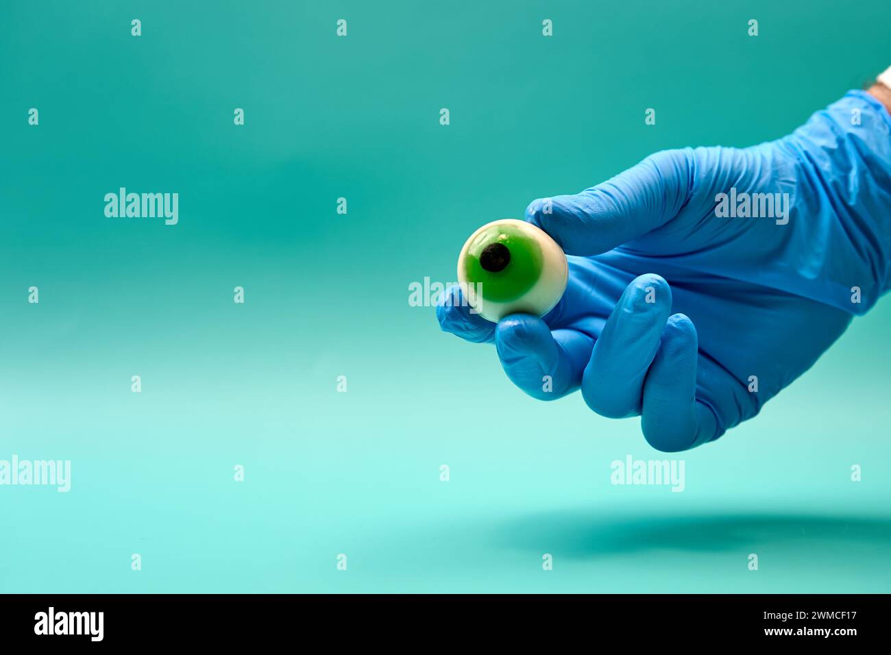 Hand of unrecognizable eye doctor in blue latex glove showing green artificial eyeball against mint green background Stock Photo