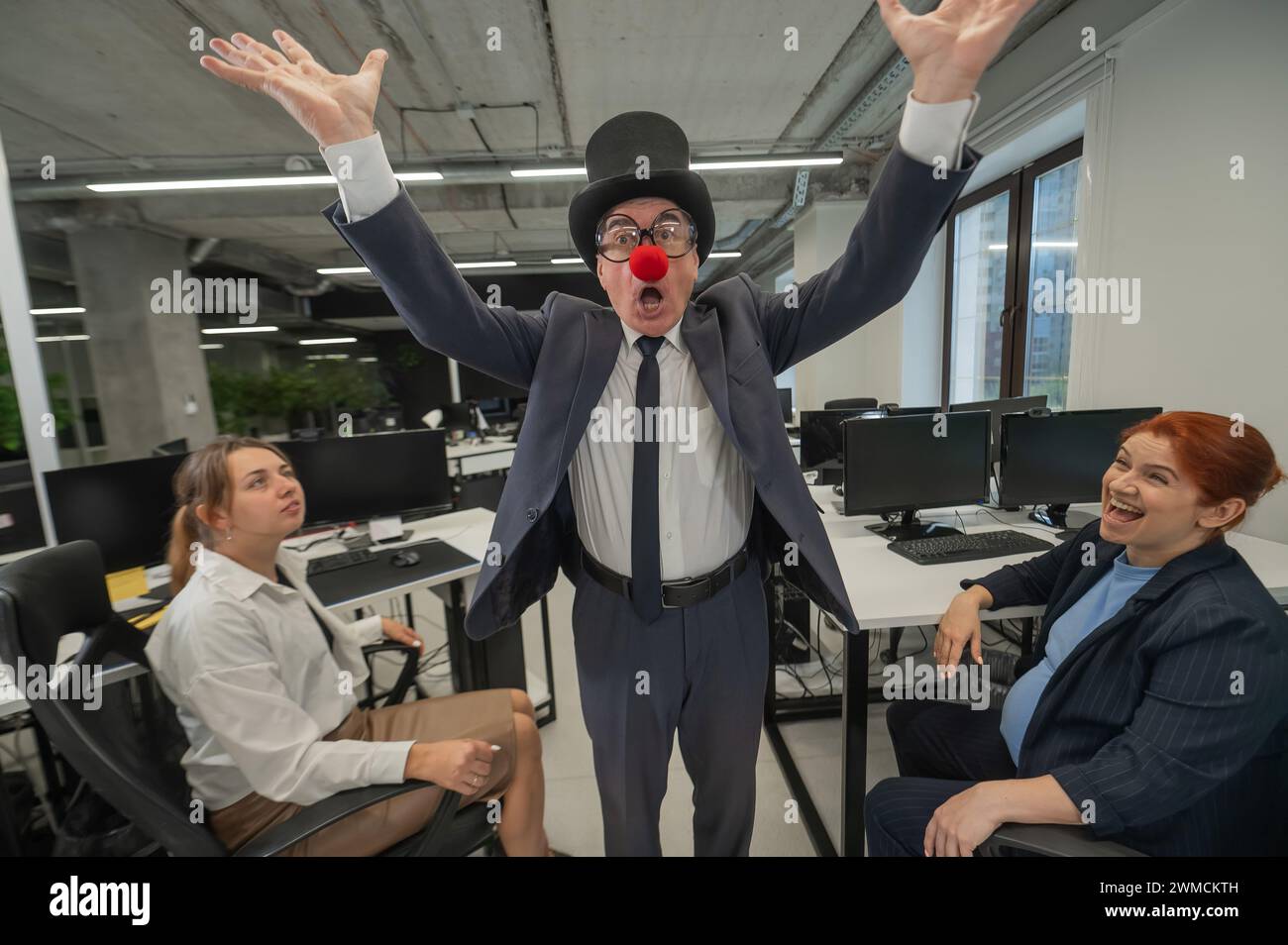 An elderly Caucasian man in a clown costume amuses two Caucasian women in the office.  Stock Photo