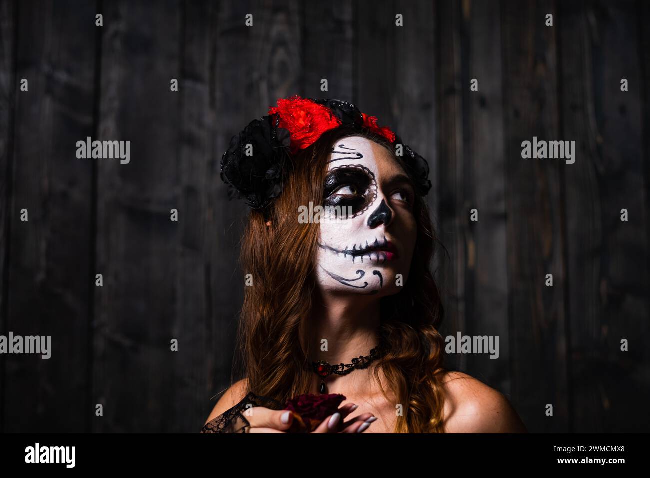 A pensive woman adorned with skull makeup gazes upwards, against a rustic wooden backdrop. Her floral headpiece adds a touch of elegance to the hauntingly beautiful scene. Stock Photo