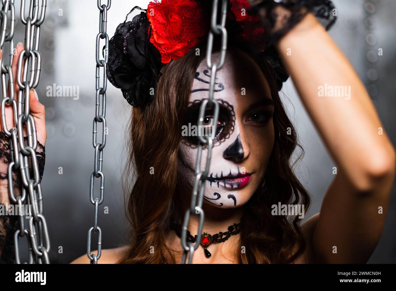 A woman with striking skull makeup peers through hanging chains, her eyes telling a story of mystique. The red and black floral headpiece contrasts with the industrial setting. Stock Photo