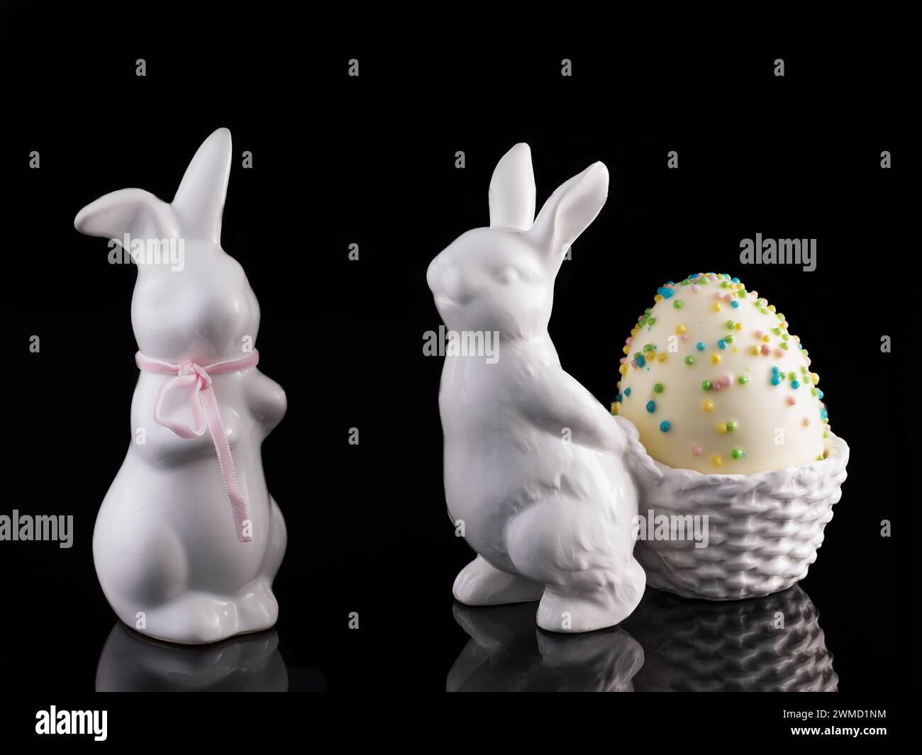 Two decorative white Poezelan Easter bunnies on a black, reflective base with a foam Easter egg Stock Photo
