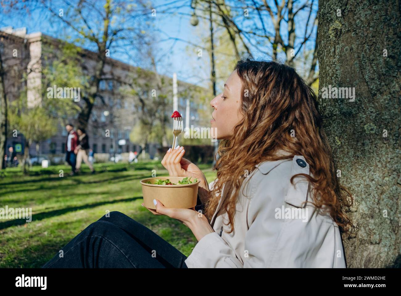 A young woman savors a bite of strawberry atop her salad, sitting serenely against a tree in a bustling city park. The moment captures the blend of urban life and the quest for wellness. Stock Photo