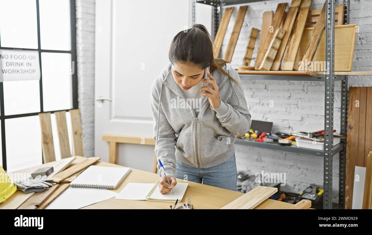Hispanic woman multitasking in a carpentry workshop, talking on the phone while taking notes. Stock Photo