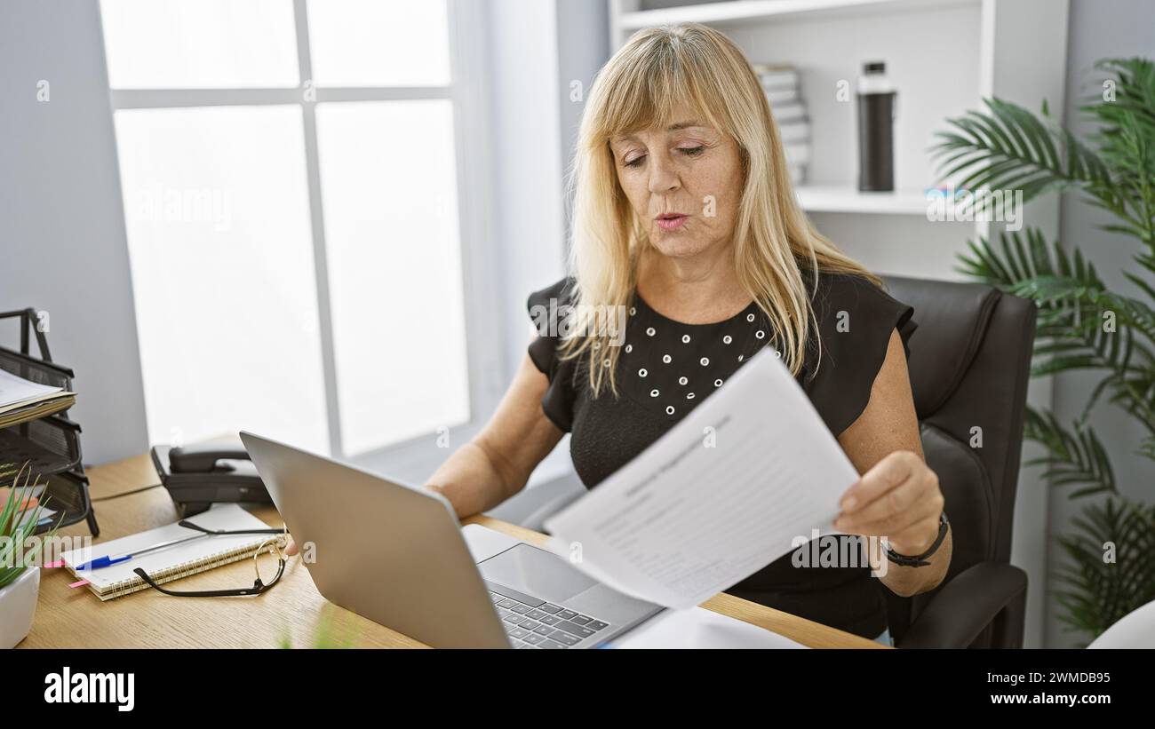 Beautiful, middle age blonde woman concentrating as she reads through work documents on her laptop in the buzzing background of a lively office enviro Stock Photo