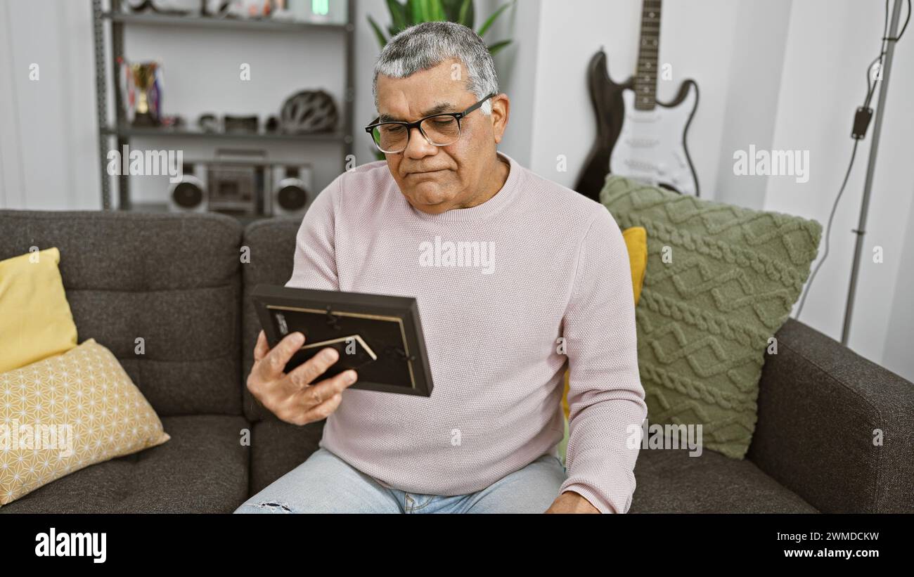 Mature man in glasses holding frame with thoughtful expression, sitting on couch in cozy living room. Stock Photo