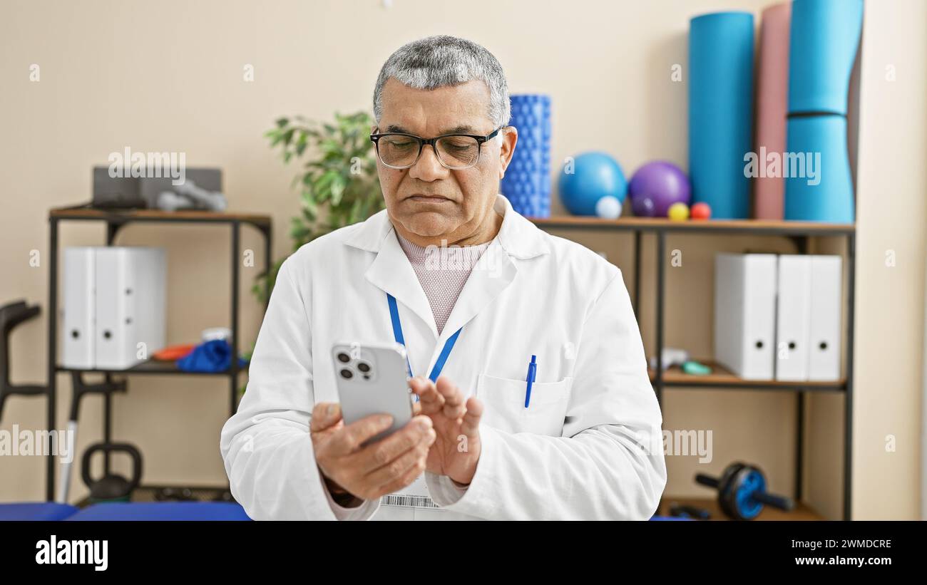 A mature man in a white lab coat using a smartphone in a rehab clinic's interior, with exercise equipment in the background. Stock Photo