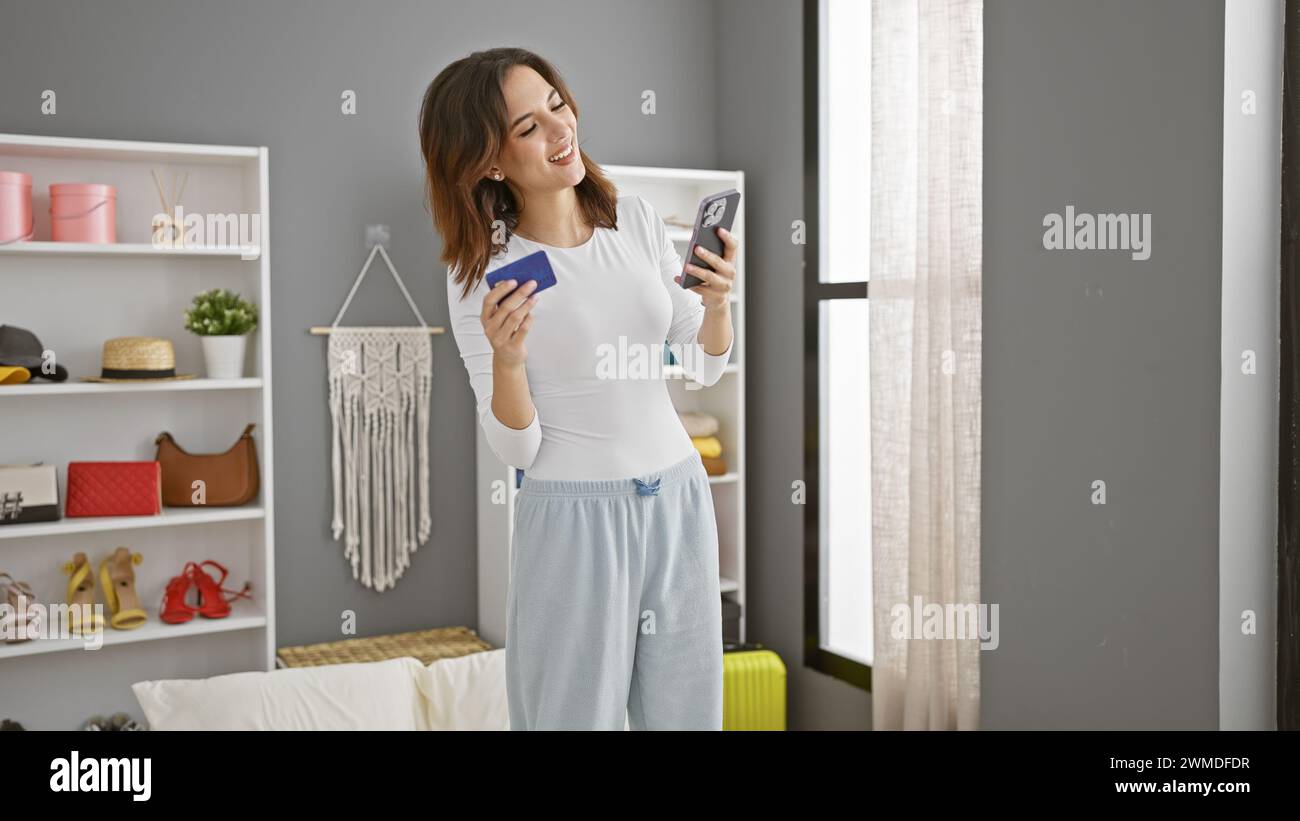 Smiling woman holding credit card and smartphone in a modern bedroom, shopping online. Stock Photo