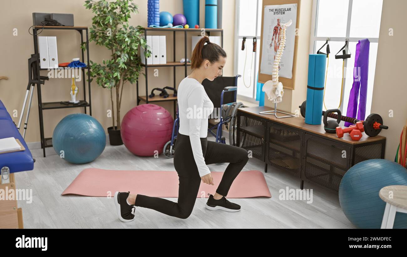 A young, hispanic woman exercising indoors at a gym with equipment like stability balls, weights, and resistance bands in the background. Stock Photo