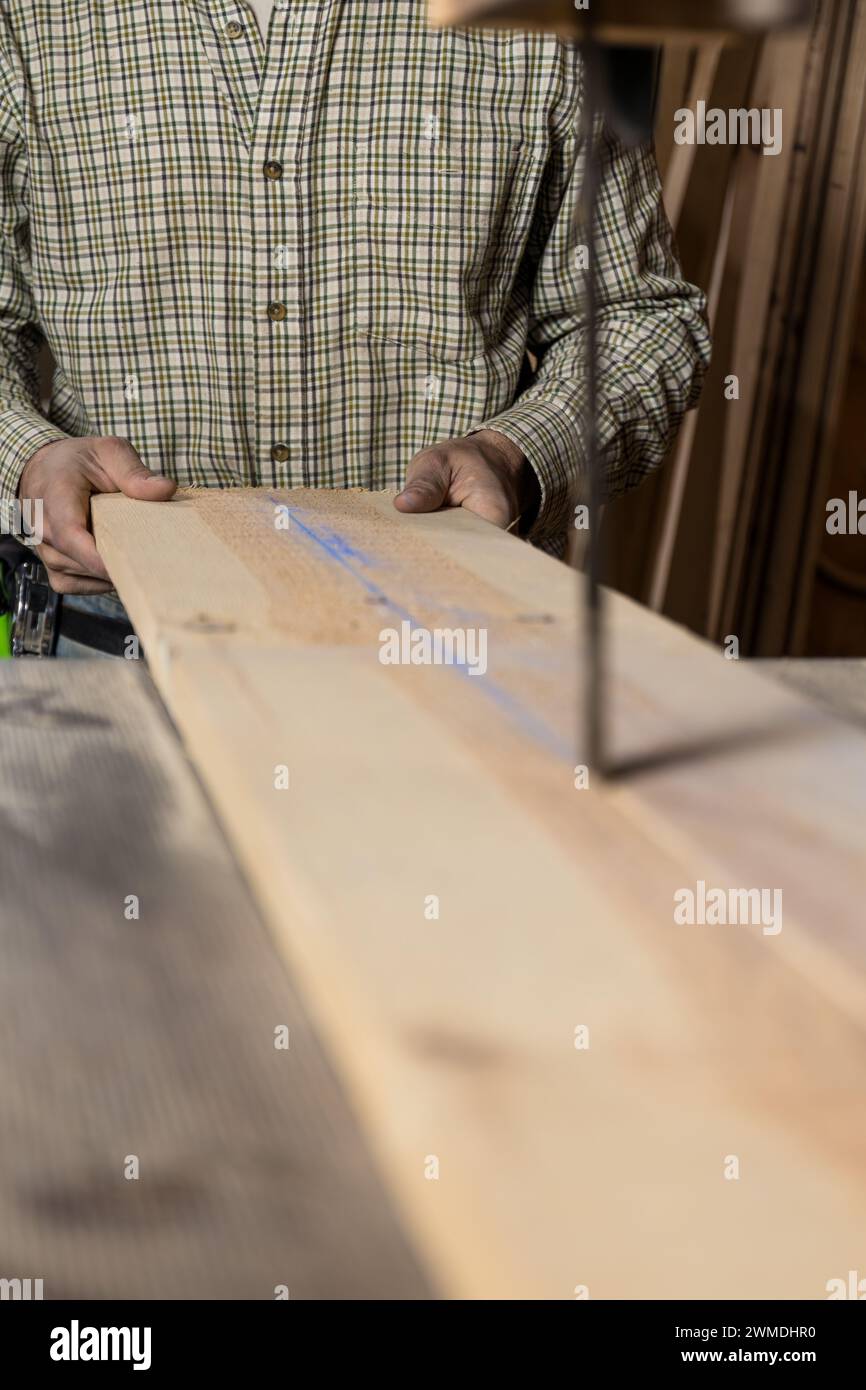 Vertical photo hands of a carpenter man mid adult caucasian carefully align a wooden plank to a bandsaw blade, with focus on the marked guideline for Stock Photo