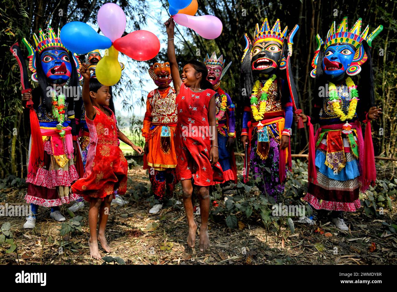 Children seen playing with balloons in front of the Masked dancers of Gomira dance troupe at a village near Raiganj. Gomira is a masked dance form. The word ‘Gomira' has been derived from the colloquial form of the word ‘Gram-Chandi' or the female deity who is the protective force of the village. The exact origin of the dance form is not traceable and the knowledge has been lost over time. Gomira dance is a rural dance form mainly practiced in the Dinajpur district of West Bengal. The Gomira dances are organized to appease the deity to usher in the 'good forces' and drive out the 'evil forces' Stock Photo