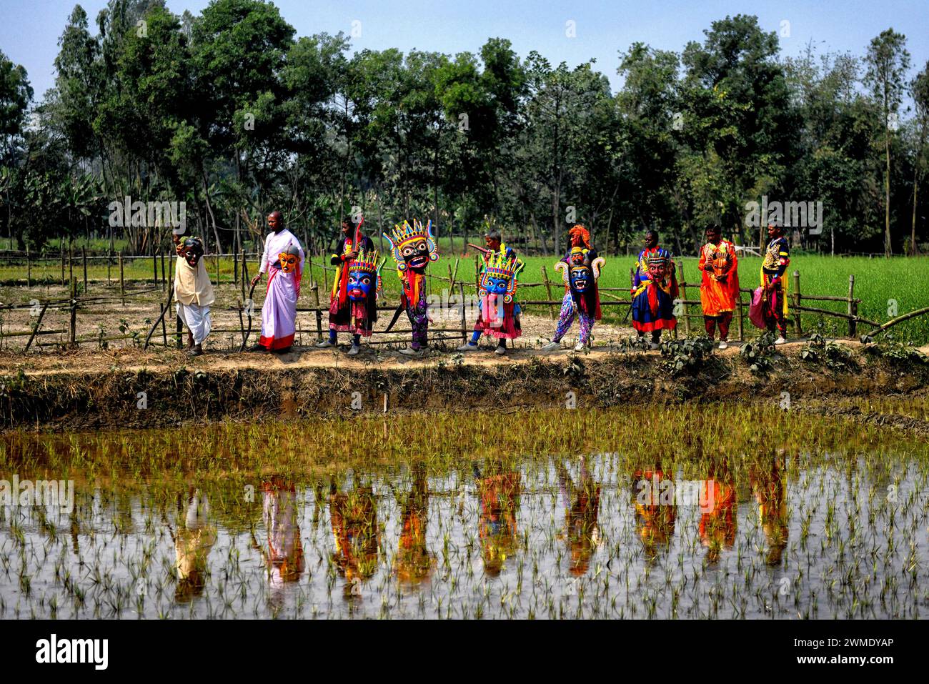 Masked dancers of Gomira dance troupe seen walking beside a water filled paddy field at a village near Raiganj. Gomira is a masked dance form. The word ‘Gomira' has been derived from the colloquial form of the word ‘Gram-Chandi' or the female deity who is the protective force of the village. The exact origin of the dance form is not traceable and the knowledge has been lost over time. Gomira dance is a rural dance form mainly practiced in the Dinajpur district of West Bengal. The Gomira dances are organized to appease the deity to usher in the 'good forces' and drive out the 'evil forces'. It Stock Photo
