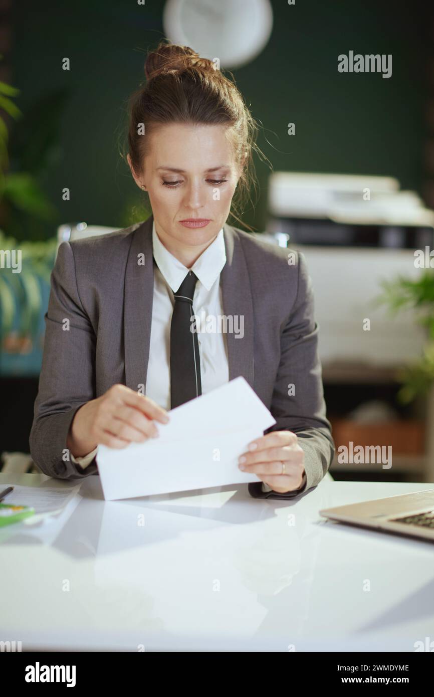 New job. pensive modern 40 years old woman employee in modern green office in grey business suit opening letter. Stock Photo
