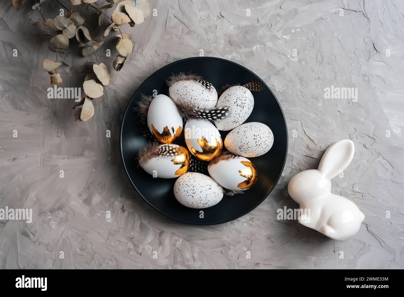 Black plate with Easter eggs, eucalyptus leaves and bunny on textured gray background. Top view, flat lay. Stock Photo