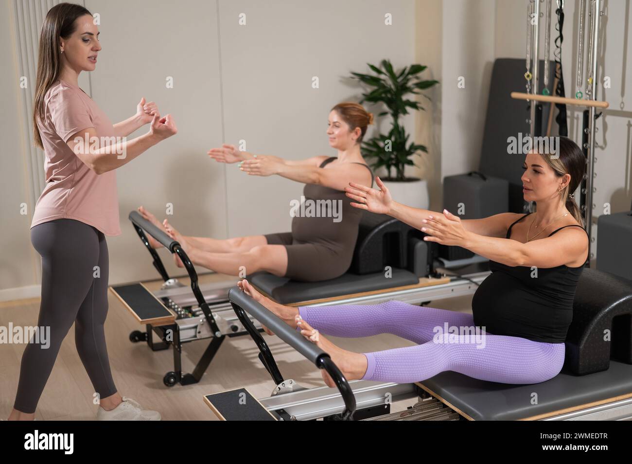 Two pregnant women are doing Pilates on a reformer. The instructor