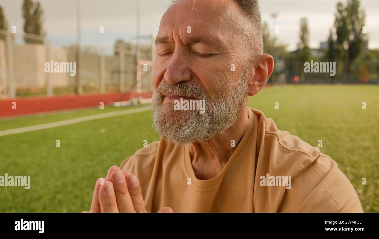 Old elderly Caucasian man stadium outside meditating on lawn sitting closed eyes smiling harmony retirement concentration wellness dreaming peaceful Stock Photo