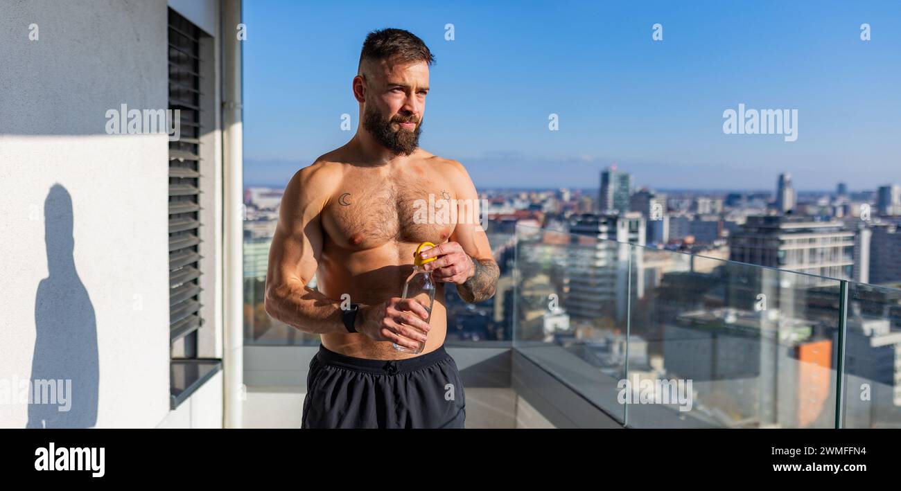 Handsome man with bare chest standing on balcony and enjoying beautiful view on the city. Relaxing after workout. Concept of selfcare and enjoying lif Stock Photo