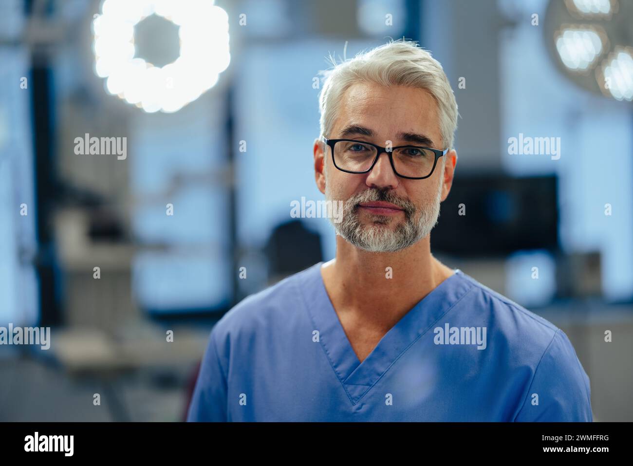 Portrait of confident ER doctor standing in hospital emergency room. Handsome doctor in blue scrubs standing in modern private clinic, looking at came Stock Photo