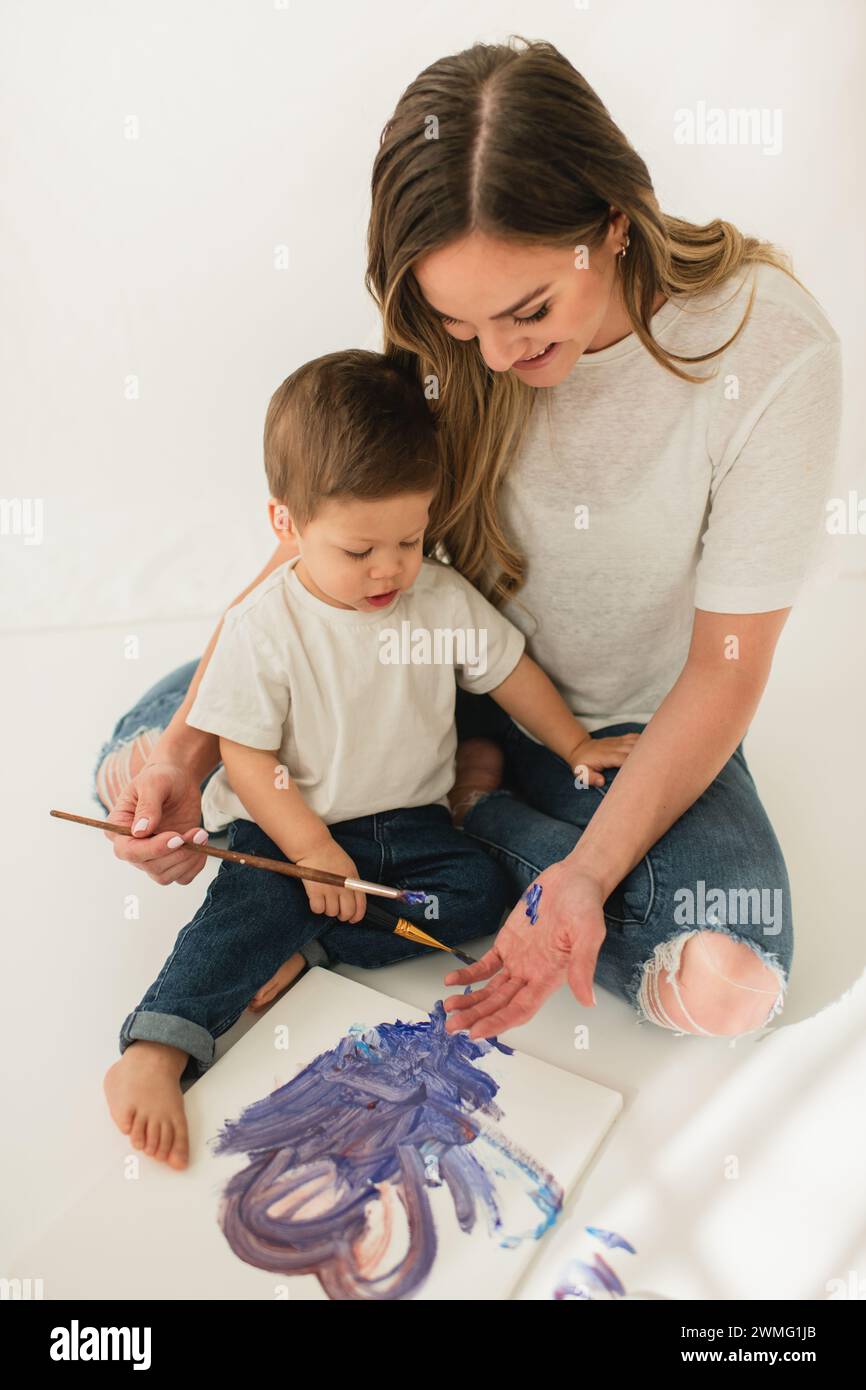Young kid painting on mom's hand using paintbrush while sitting Stock Photo