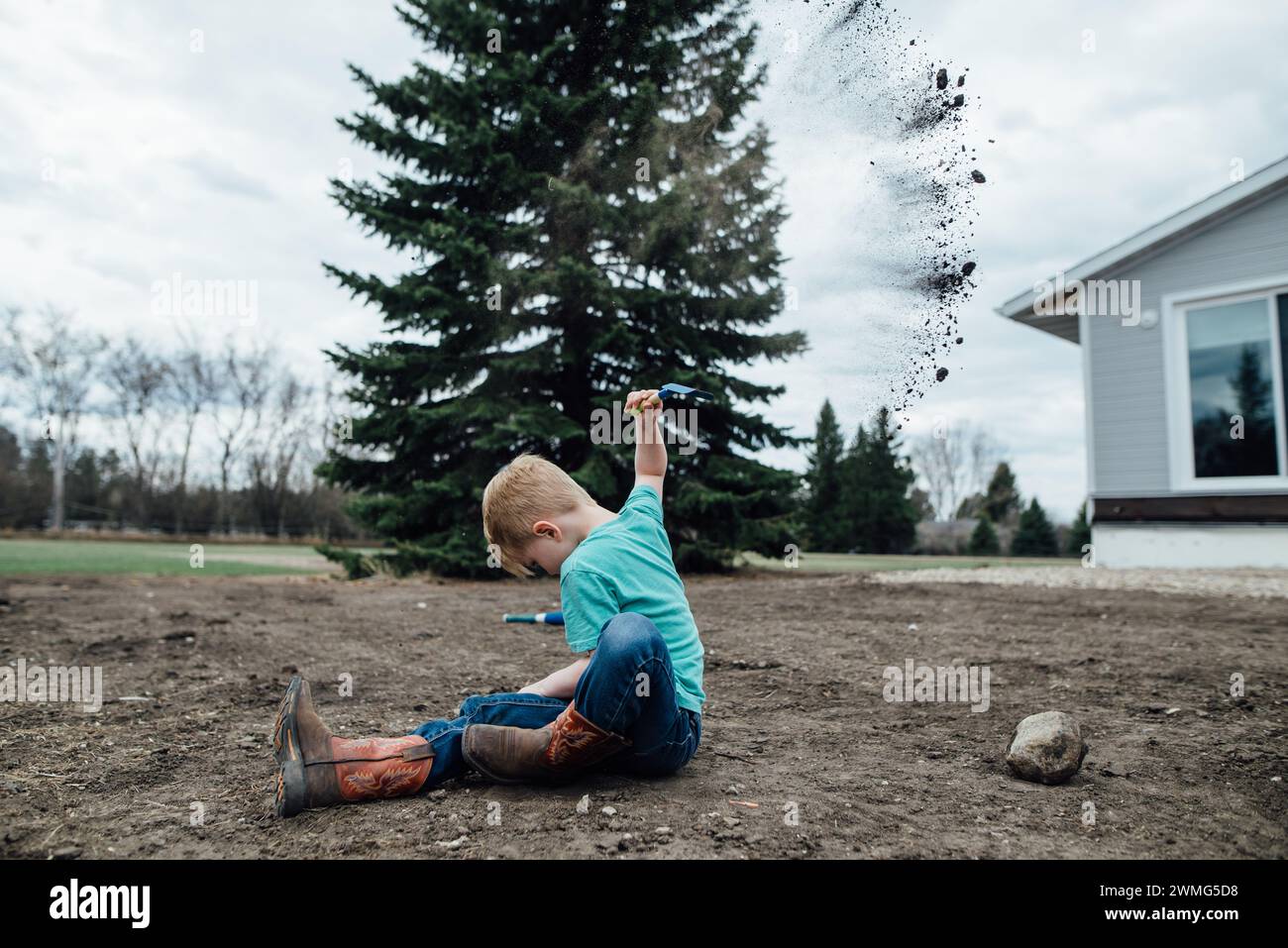 Side view of young boy sitting in yard while throwing handfuls of dirt Stock Photo