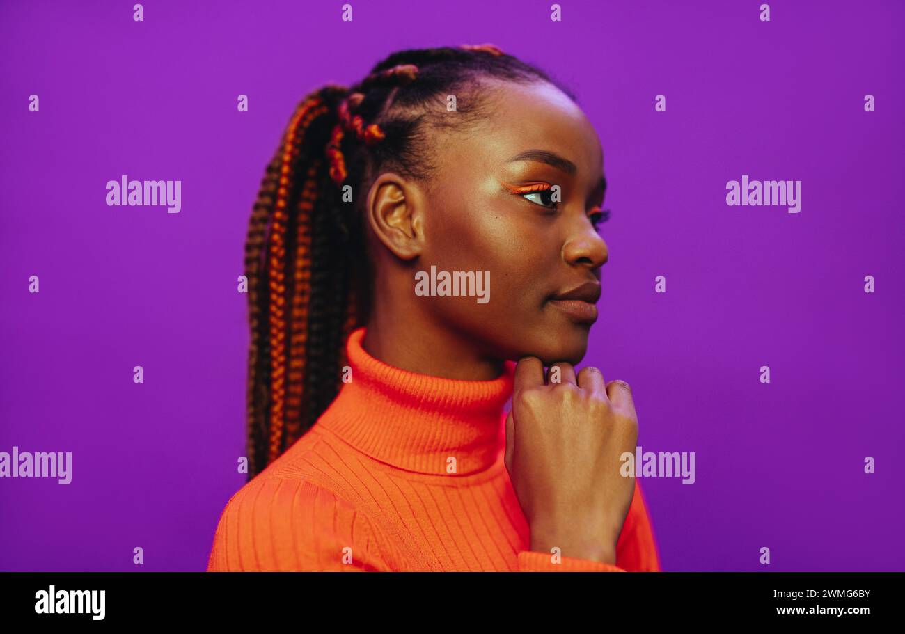 Confident gen Z woman in her 20s with stylish braids, vibrant makeup, and casual clothing poses against a purple background with close-up portrait. Stock Photo