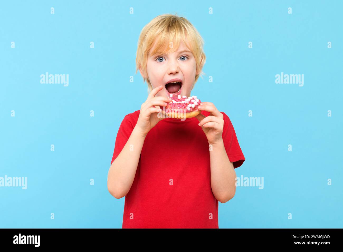 Cute little boy with blond hair and freckles eating a glazed donut. Children and sugary junk food concept. Boy holding colorful donuts, eating junk un Stock Photo