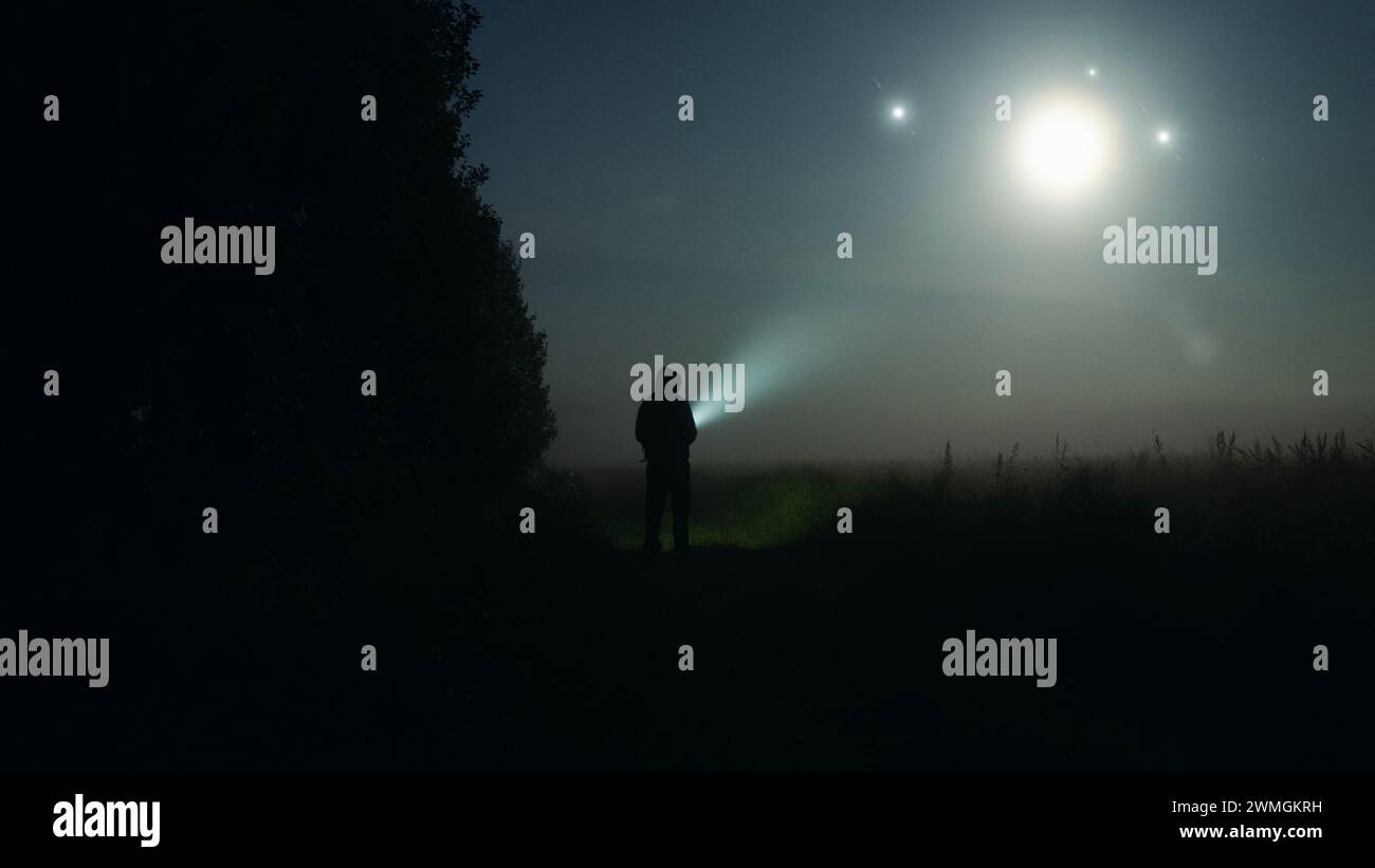 A mysterious figure with torch looking at glowing UFO lights in the sky.. On a spooky misty night in the countryside. Stock Photo