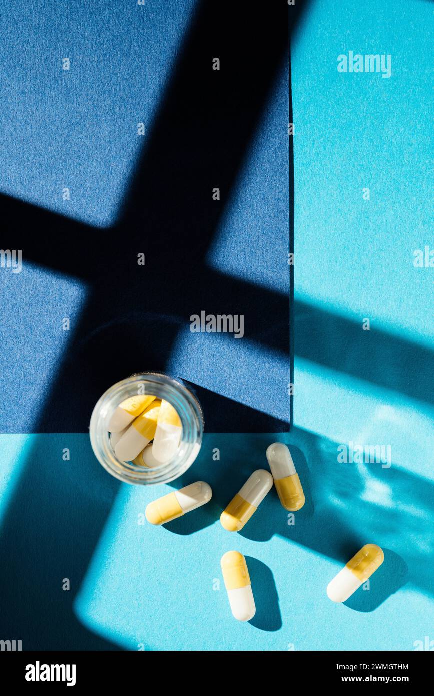 White and yellow capsule pills in plastic container on blue background under direct light with shadow effect Stock Photo