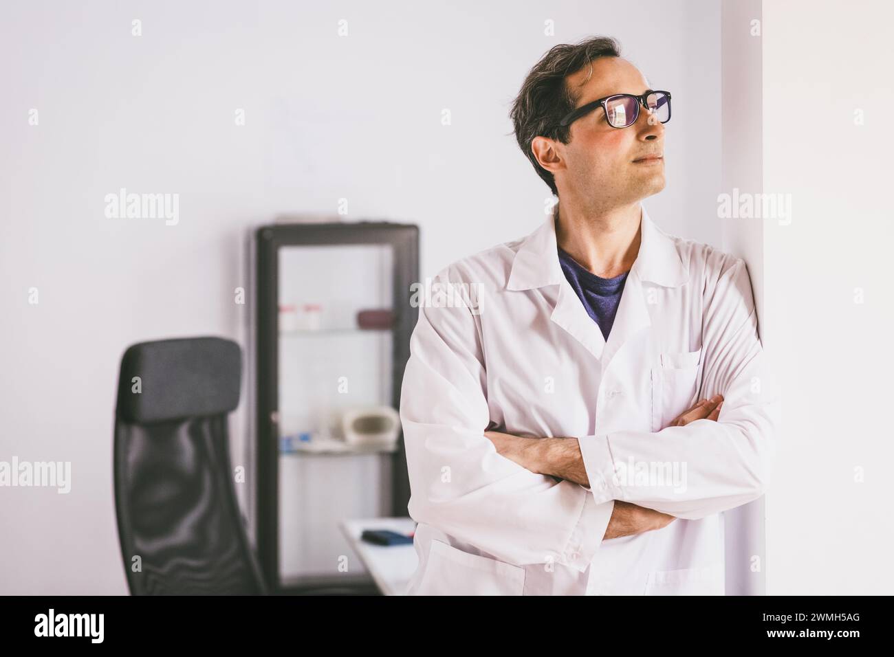 An adult male Caucasian scientist in glasses and a white coat stands and looks forward against the backdrop of a laboratory. Stock Photo
