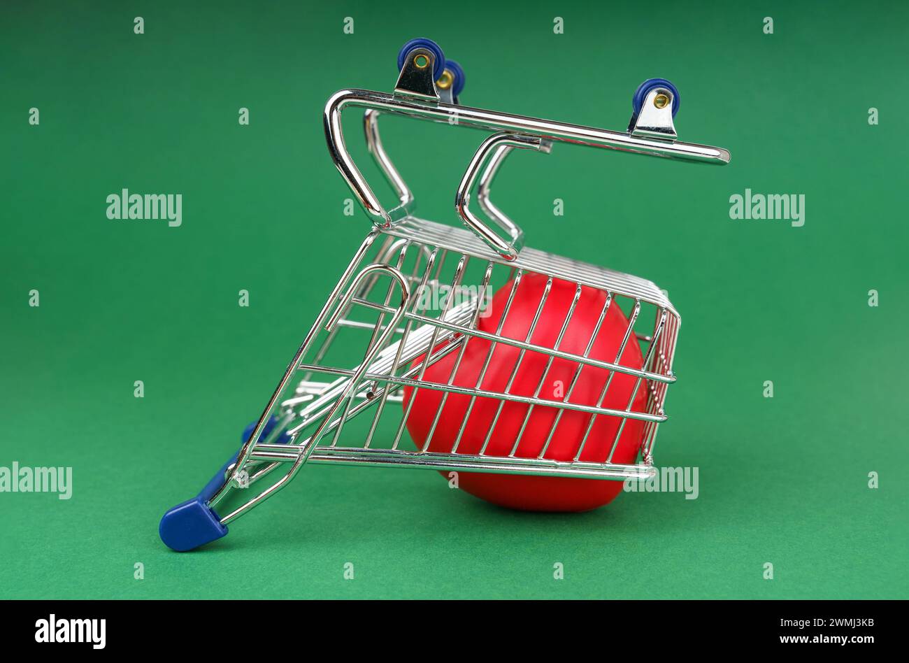On a solid green background sits a small metal shopping cart with blue handles containing a red heart. The cart stands upside down, simulating disappo Stock Photo