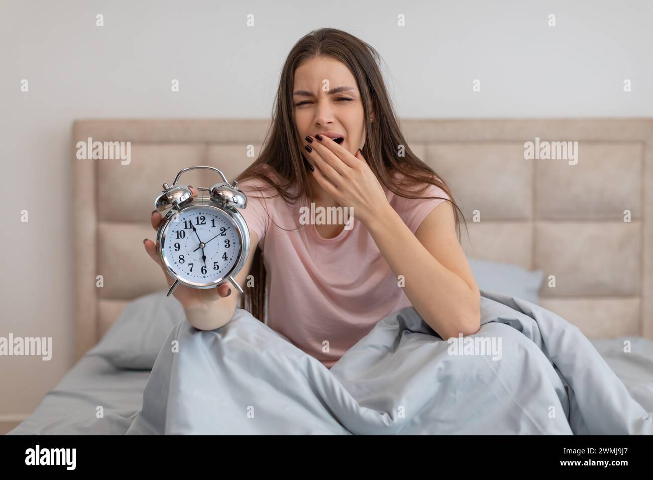 Sleepy woman in bed holding an alarm clock, yawning with tiredness Stock Photo
