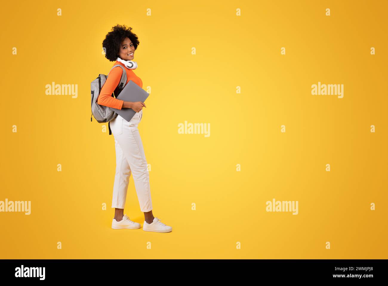 Confident young student in a vibrant orange turtleneck and white pants looks back with a smile Stock Photo