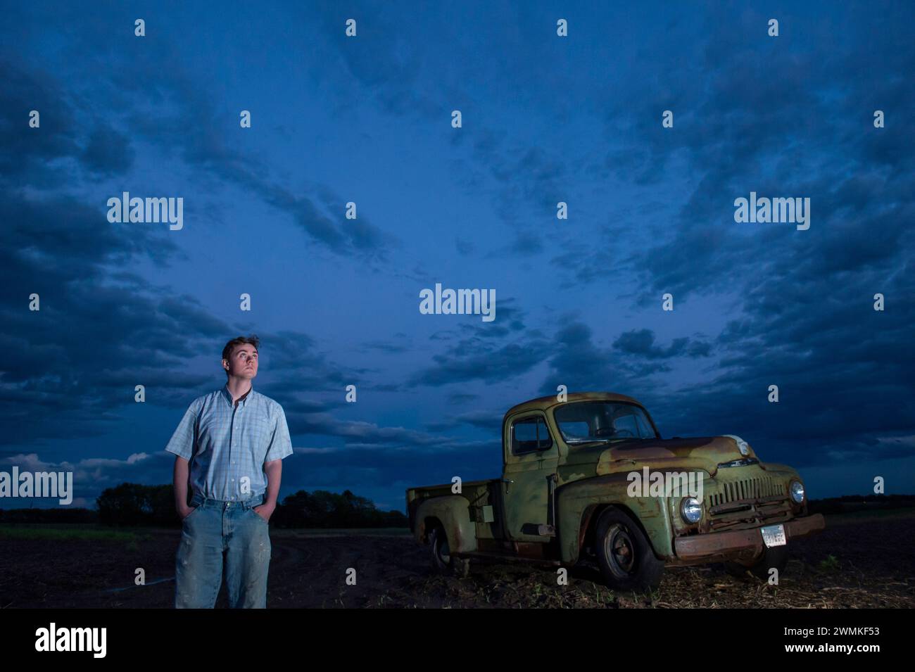 Young man looks at the gathering storm clouds in the sky near a 1951 International Harvester pickup truck on a farm field Stock Photo