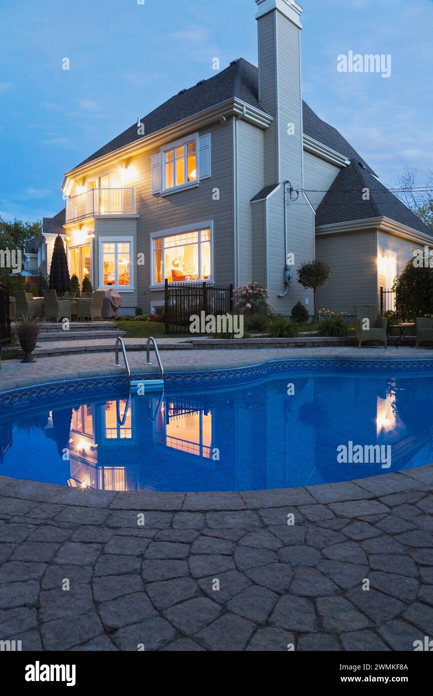 Modern two story tan clapboard cladded home with landscaped backyard and In-ground swimming pool illuminated at dusk. Stock Photo