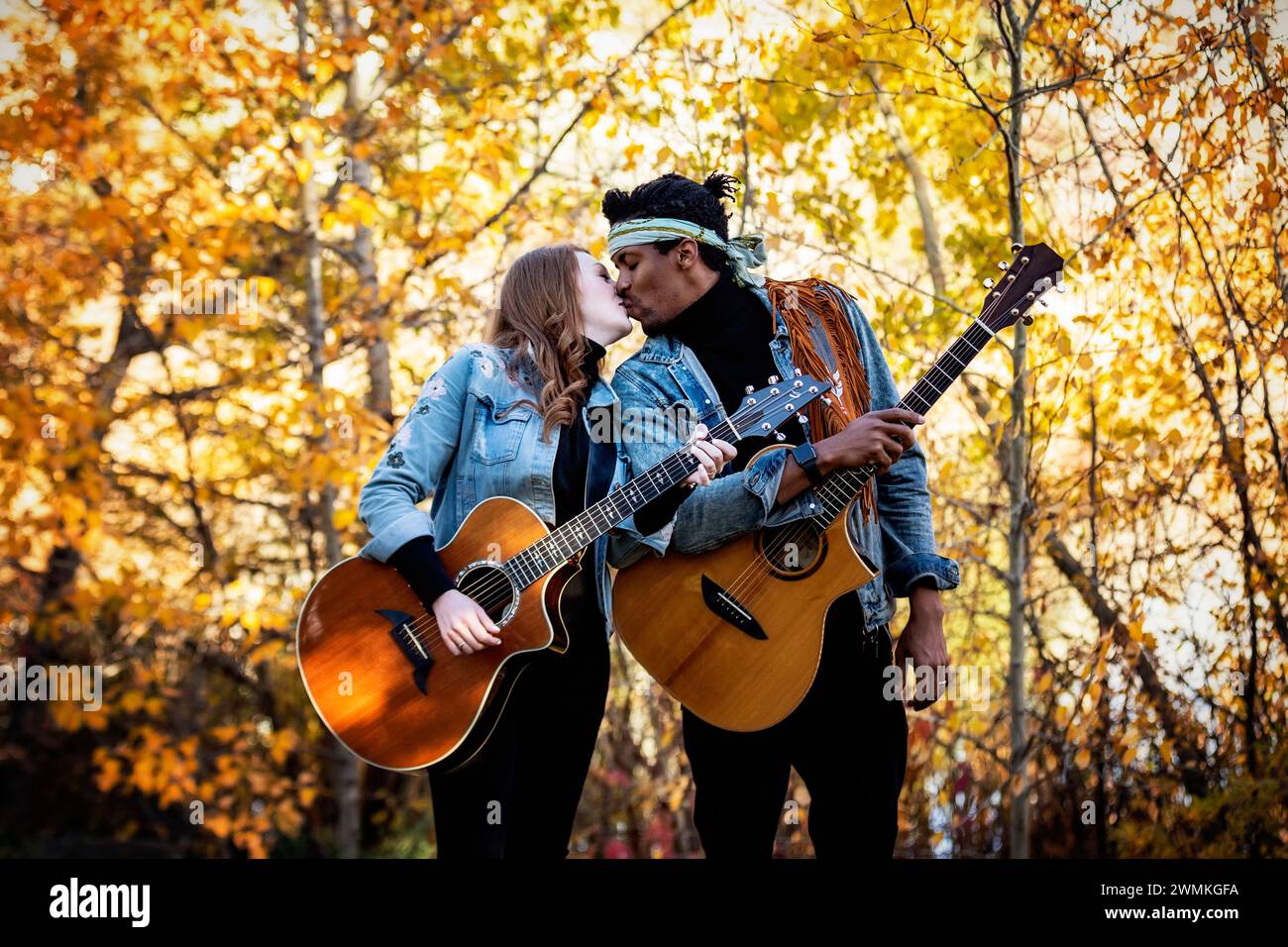 Mixed race married couple standing with guitars, kissing each other during a fall family outing in a city park, spending quality time together Stock Photo
