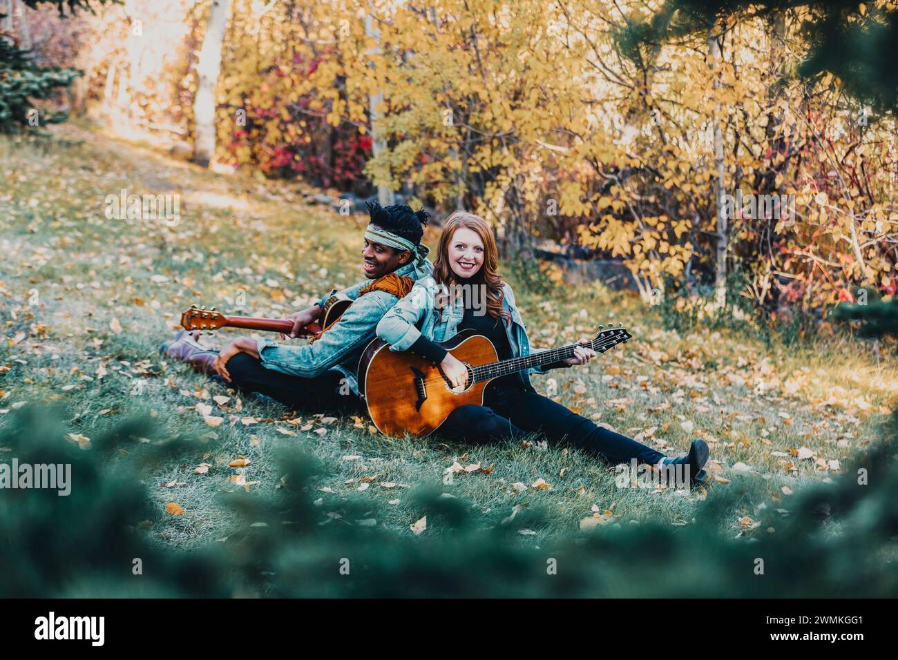 Framed view of a mixed race married couple sitting on the grass back to back playing guitars, smiling and posing for the camera during a fall famil... Stock Photo