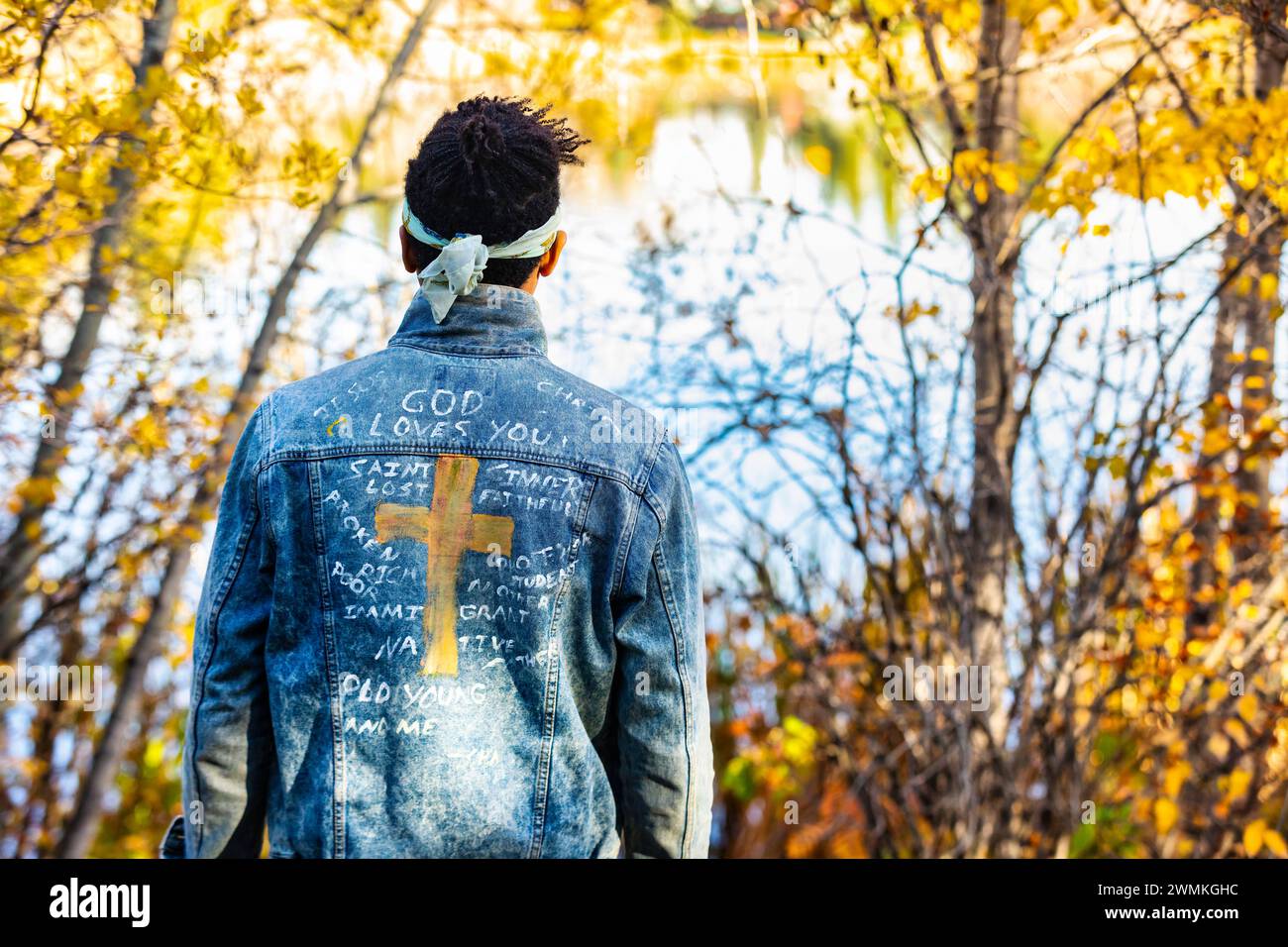 Close-up view from behind of a man wearing a jean jacket with hand-written Christian references during a fall outing in a city park, standing by a ... Stock Photo