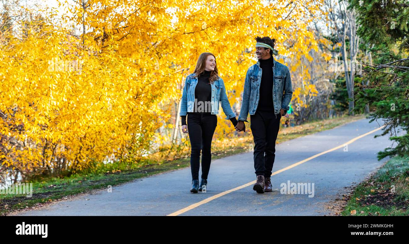 A mixed race married couple smiling at each other, walking down a road in a city park during a fall family outing, spending quality time together Stock Photo