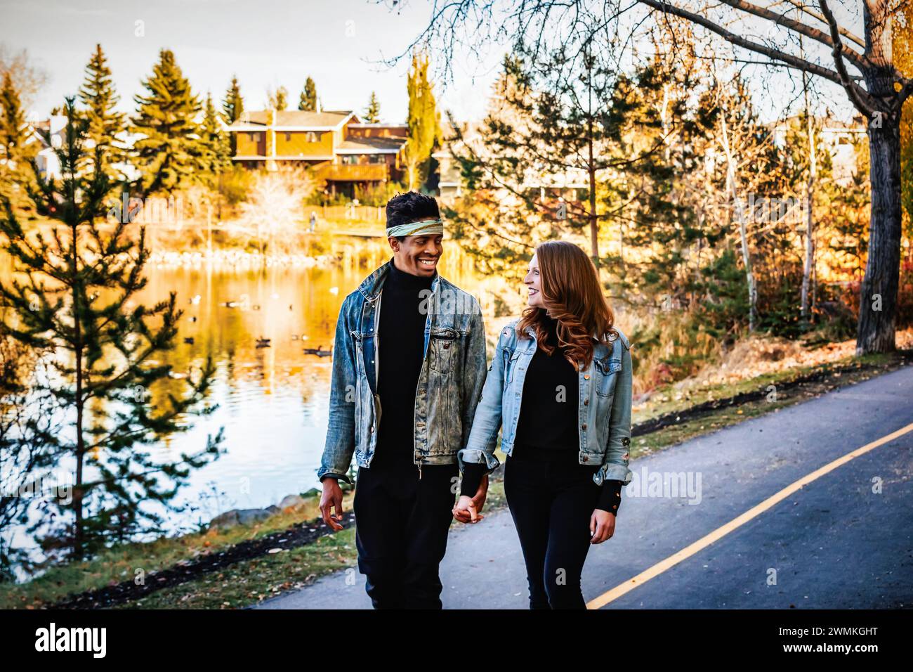 A mixed race married couple holding hands and smiling at each other, walking down a road in a city park during a fall family outing, spending quali... Stock Photo