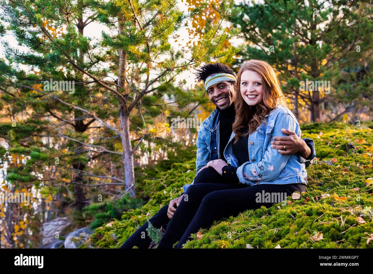 Portrait of a mixed race couple embracing and smiling at the camera, sitting on a grassy hillside, spending quality time together during a fall fam... Stock Photo