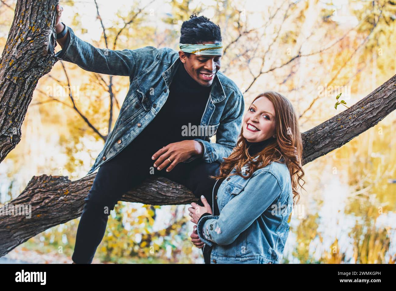 Close-up portrait of a mixed race couple smiling while resting on a tree branch, spending quality time together during a fall family outing in a ci... Stock Photo
