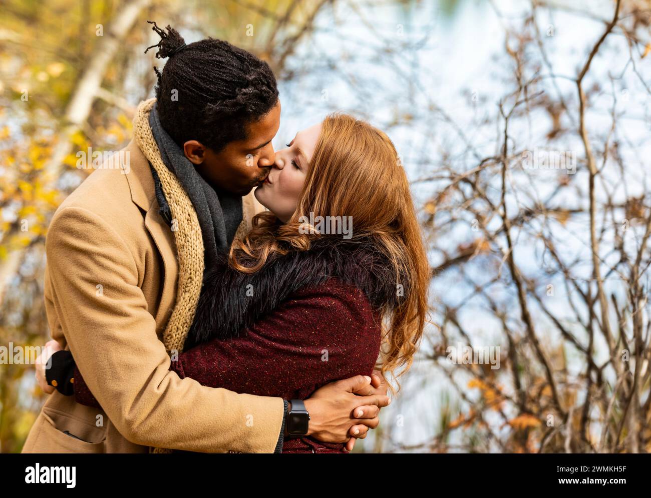 A mixed race couple standing by the shore of a lake hugging and kissing each other while spending quality time together during a fall family outing... Stock Photo