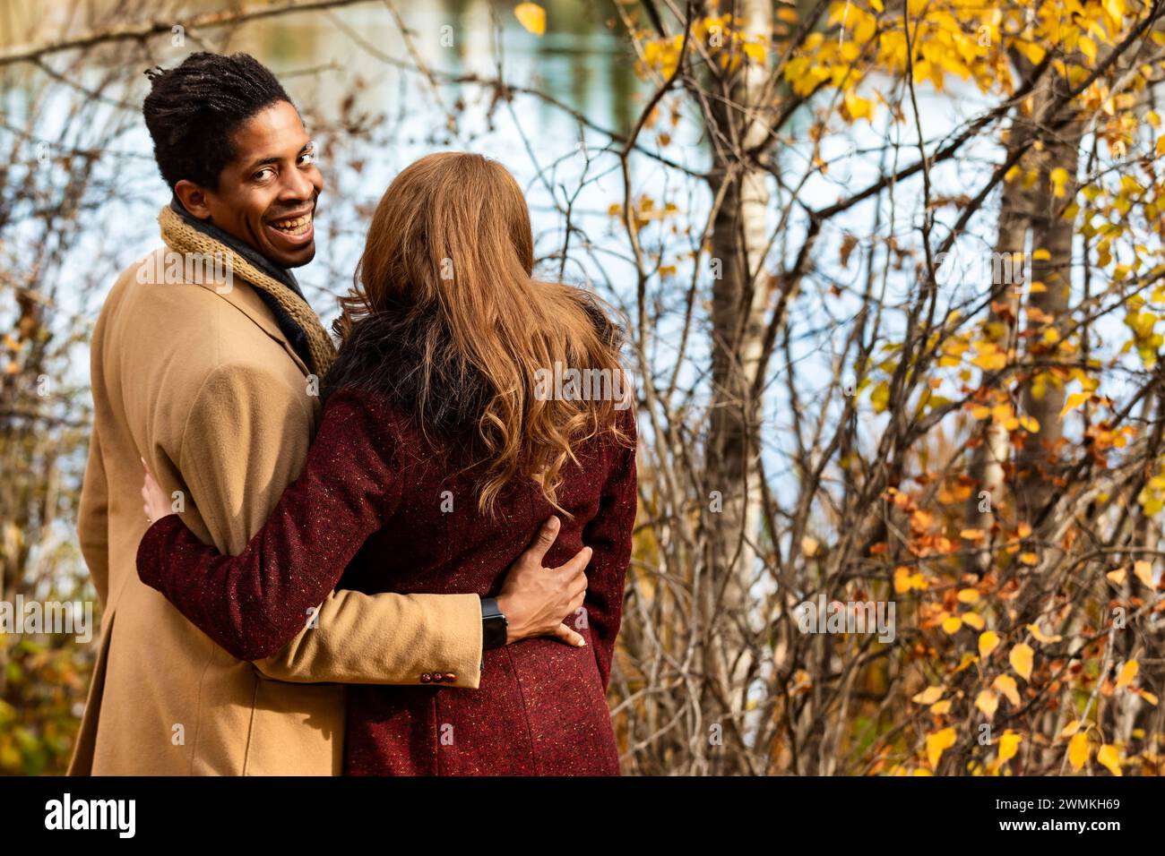 View taken from behind of a mixed race couple standing by the shore of a lake hugging each other with husband looking back and smiling at the camer... Stock Photo