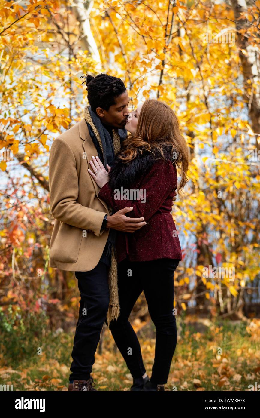 A mixed race couple standing by the shore of a lake hugging and kissing each other while spending quality time together during a fall family outing... Stock Photo