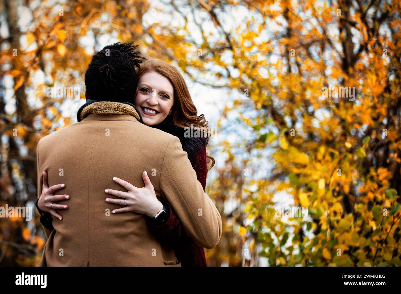 View taken from behind of a mixed race married couple hugging each other with wife smiling at the camera, while spending quality time together duri... Stock Photo