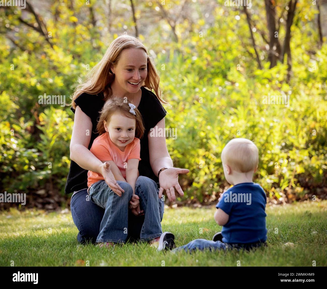 Mother throwing a ball with her young son who has Down Syndrome, and her preschooler daughter, in a city park during a warm fall afternoon Stock Photo