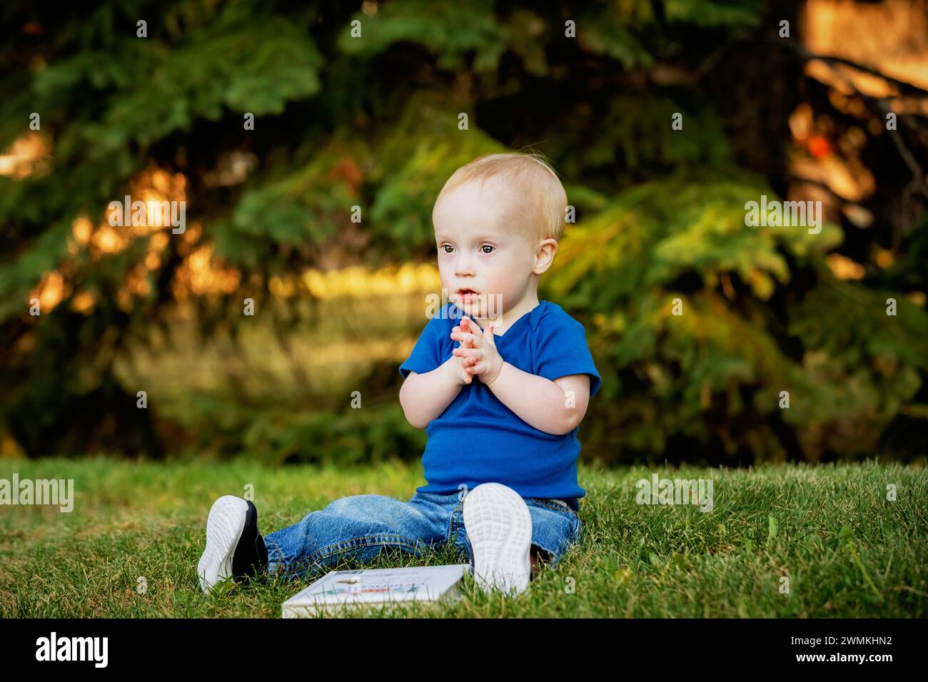 Baby boy with Down syndrome sitting on grass in a city park with a book and using sign language, during a warm fall afternoon; Leduc, Alberta, Canada Stock Photo