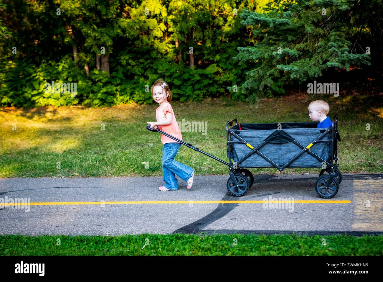 Preschooler girl pulling her younger brother who has Down syndrome in a wagon on a path at a city park during a warm fall evening Stock Photo
