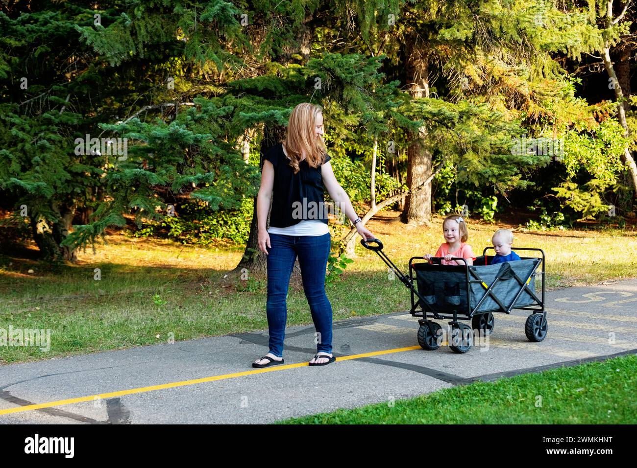 Mother pulling a wagon with her young son who has Down Syndrome and her preschooler daughter, in a city park during an autumn day Stock Photo